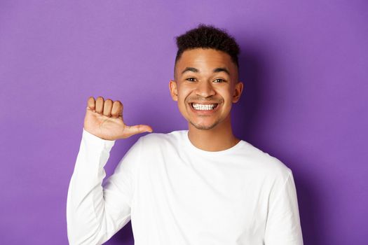 Close-up of young cheerful african-american guy, smiling and pointing at himself, standing over purple background in white sweatshirt.