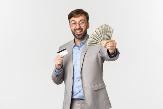 Portrait of happy successful businessman holding credit card, showing money and rejoicing, standing over white background.