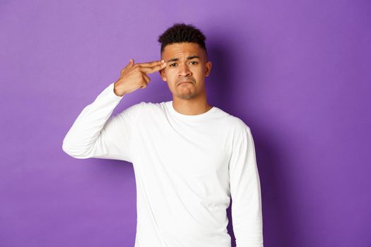 Image of sad and tired african-american male student, making finger gun sign over head and shooting himself, standing distressed against purple background.