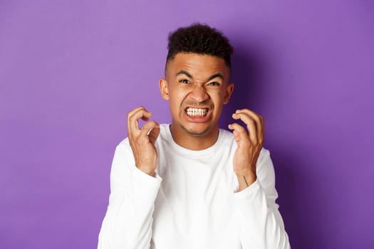 Close-up of angry and annoyed african-american man, clench fists and grimacing from scorn and hatred, standing furious over purple background.