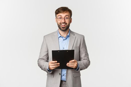 Image of confident surprised and happy businessman in grey suit and glasses, holding clipboard and looking at camera, standing over white background.