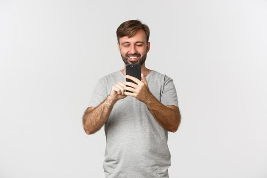 Portrait of adult man with beard, wearing gray t-shirt, taking photo on smartphone or recording a video, standing over white background.