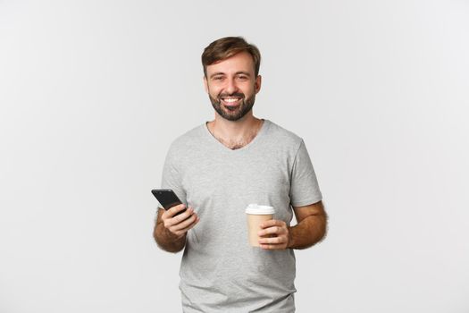 Image of modern caucasian man with beard, looking happy while drinking coffee and casually using mobile phone, standing over white background.