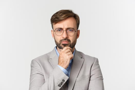 Close-up of thoughtful bearded businessman, wearing glasses and gray suit, thinking about something with serious face, standing over white background.