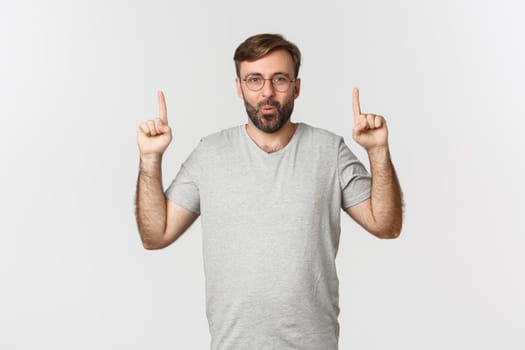 Surprised and excited bearded man smiling, pointing fingers up, showing logo, wearing gray t-shirt, standing over white background.