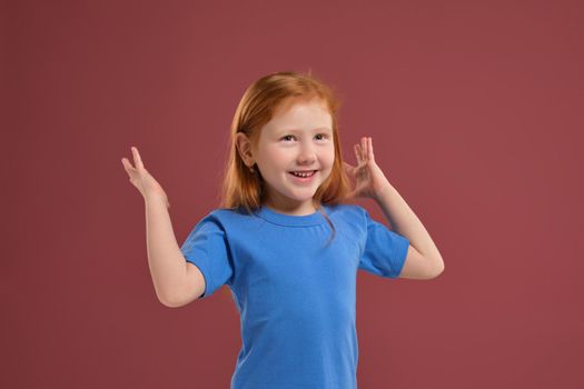 Portrait of cute redhead emotional little girl in blue t-shirt on red background. Beautiful studio portrait of female little caucasian person spreading her hands, she's so happy