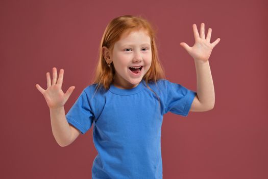 Portrait of cute redhead emotional little girl in blue t-shirt on red background. Beautiful studio portrait of female little caucasian person spreading her hands, she's so happy