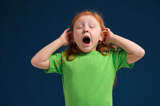 close up photo of little redhead emotional girl in green t-shirt posing before camera. She is tired and wants to sleep, yawning and stretching