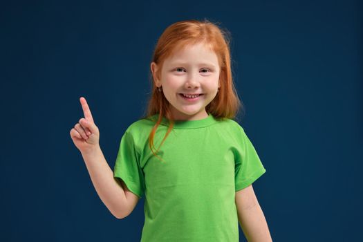 close up view of adorable happy redhead little girl in green t-shirt on blue background pointing up with her finger, smiling