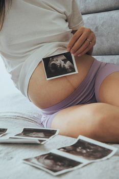Close up at young pregnant caucasian woman, future mother, sitting on comfortable bed and looking at ultrasound x ray photos of her baby in tummy.
