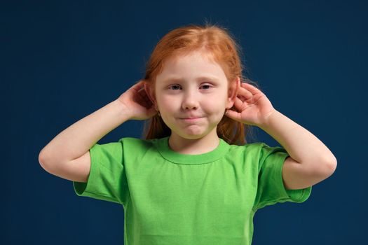Portrait of cute redhead little girl trying to hear someone putting her hand on the ear, blue background, green t-shirt