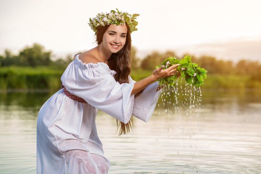 Beautiful black haired girl in white vintage dress and wreath of flowers standing in water of lake. Fairytale story. Warm art work. Sun flare