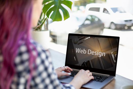 Cropped shot view of young woman working on laptop computer while sitting in modern cafe bar interior, pink hair female freelancer using net-book for remote job during morning breakfast in coffee shop. Design. Web design