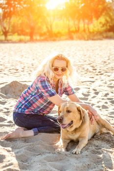 Portrait of young beautiful woman in sunglasses sitting on sand beach with golden retriever dog. Girl with dog by sea. Happiness and friendship. Pet and woman. Woman playing with dog on sea shore. Sun flare