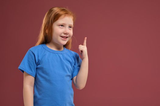 Portrait of cute redhead emotional little girl in blu t-shirt on red background. I have an idea Eureka bingo