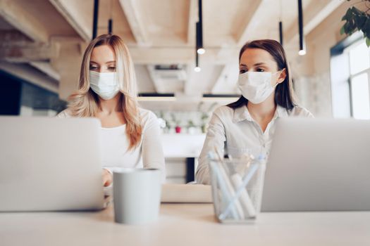 Two female colleagues working in office together wearing medical masks, coronavirus prevention