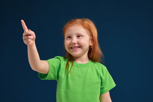 close up view of adorable happy redhead little girl in green t-shirt on blue background pointing up with her finger, smiling
