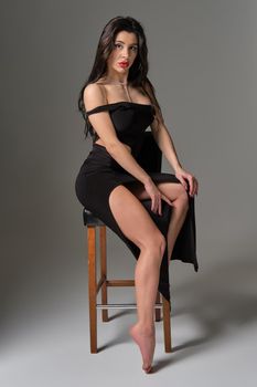 Photo of young attractive brunette caucasian sexy woman sits on chair wearing erotic cocktail black dress. Gray background, studio shot high quality image