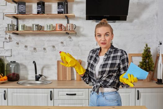 Housekeeper concept. Professional young woman wearing rubber protective yellow gloves holding bottle cleaners in the kitchen. Housewife hold bottle spray work clean at home.
