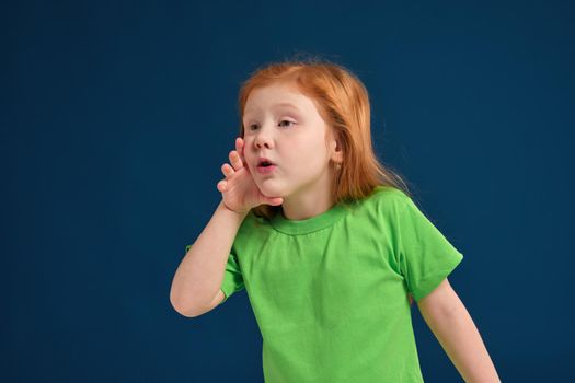 Portrait of a small pretty redhead girl standing and calling someone holding her hand near her mouth wearing a green t-shirt ,blue background