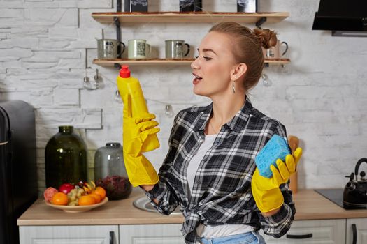 Housekeeper concept. Professional young woman wearing rubber protective yellow gloves holding bottle cleaners in the kitchen. Housewife hold bottle spray work clean at home.