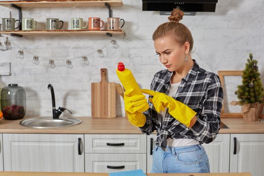 Housekeeper concept. Professional young woman wearing rubber protective yellow gloves holding bottle cleaners in the kitchen. Housewife hold bottle spray work clean at home.