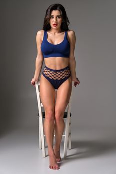 Full length studio shot of a glamour brunette lady in a sexy lingerie posing against a white chair on a gray background. Seductive model with a bright make-up and in a blue underwear. Fashionable lifestyle, sensuality and beauty concept.