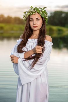 Beautiful black haired girl in white vintage dress and wreath of flowers standing in water of lake. Fairytale story. Warm art work. Sun flare