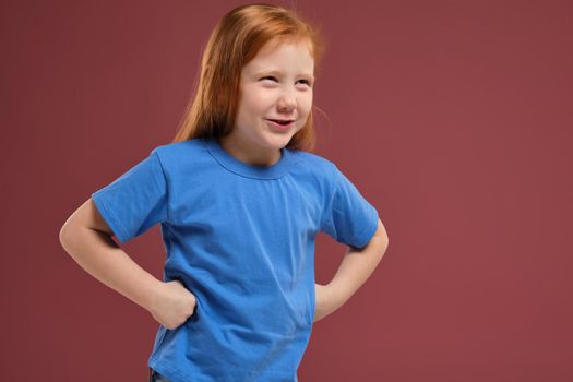 close-up portrait of cute redhead emotional little girl in blue t-shirt on red background. She is about to get nervous, angry with someone