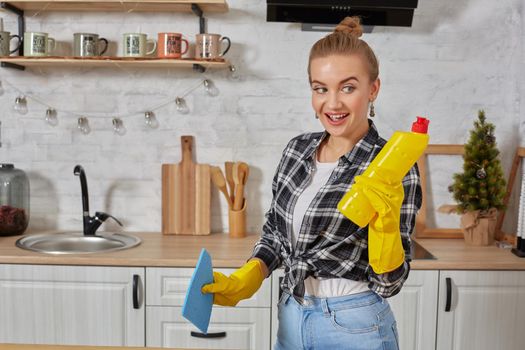 Housekeeper concept. Professional young woman wearing rubber protective yellow gloves holding bottle cleaners in the kitchen. Housewife hold bottle spray work clean at home.