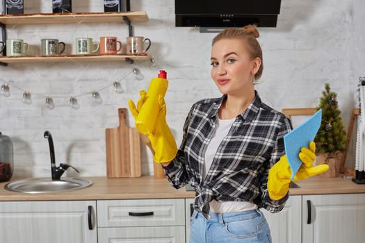 Housekeeper concept. Professional young woman wearing rubber protective yellow gloves holding bottle cleaners in the kitchen. Housewife hold bottle spray work clean at home.