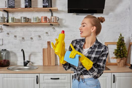 Housekeeper concept. Professional young woman wearing rubber protective yellow gloves holding bottle cleaners in the kitchen. Housewife hold bottle spray work clean at home.