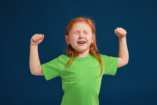 close up photo of little redhead emotional girl in green t-shirt posing before camera.She is tired and wants to sleep, yawning and stretching
