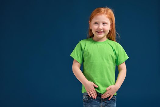 Portrait of cute redhead emotional little girl in green t-shirt on blue background. Beautiful studio portrait of female little caucasian person