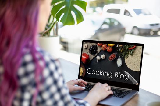 Back view of young pink hair woman watching blog video about healthy food on laptop while sitting in cafe