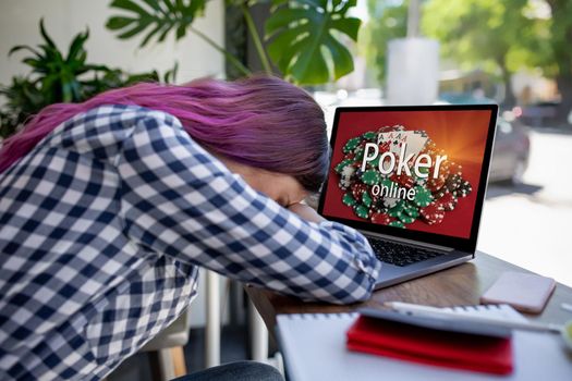 Side view of a young pink hair woman keyboarding on laptop computer with poker online on a screen while sitting in cafe. Play online concept. Screen graphics are made up. Emotions. Loss