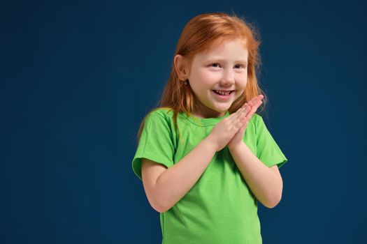 Sneaky cute redhead little girl rub her hands with grin on her face, blue background, green t-shirt