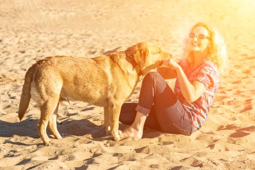 Portrait of young beautiful woman in sunglasses sitting on sand beach hugging golden retriever dog. Girl with dog by sea. Happiness and friendship. Pet and woman. woman playing with dog on sea shore. Sun flare