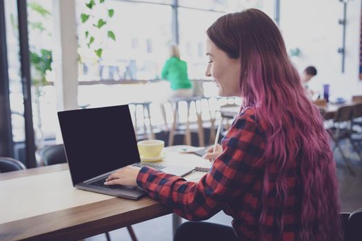 Rear shot of female student learning online via laptop before her lectures. Woman typing on notebook with blank screen with copy space for your text or advertising content, sitting at wooden table