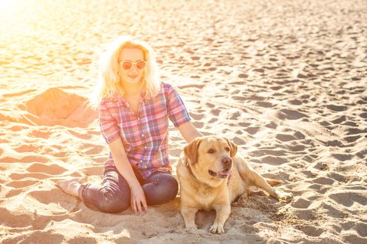 Portrait of young beautiful woman in sunglasses sitting on sand beach with golden retriever dog. Girl with dog by sea. Happiness and friendship. Pet and woman. Woman playing with dog on sea shore. Sun flare