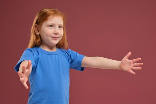 Portrait of cute redhead emotional little girl in blue t-shirt on red background. Beautiful studio portrait of female little caucasian person spreading her hands, she's so happy