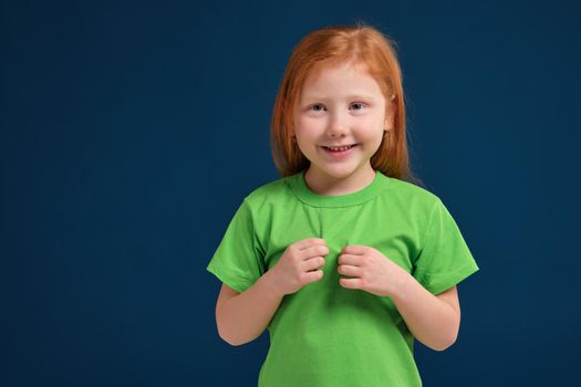 Portrait of cute redhead emotional little girl in green t-shirt on blue background. Beautiful studio portrait of female little caucasian person. She looks so happy and shy a little bit