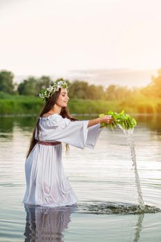 Beautiful black haired girl in white vintage dress and wreath of flowers standing in water of lake. Fairytale story. Warm art work. Sun flare