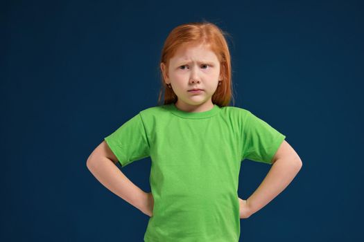 Portrait of angry offended displeased redhead little girl, holding hands on hips, looking from under forehead
