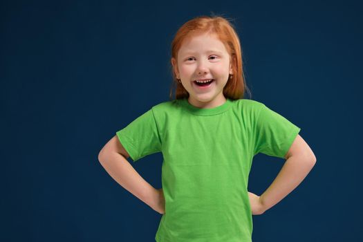 Close up portrait of happy young redhead little girl smiling at camera and holding hands on hips