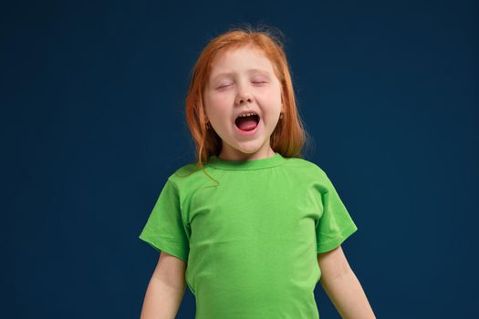 close up photo of little redhead emotional girl in green t-shirt posing before camera. She is tired and wants to sleep, yawning and stretching