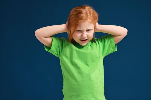 Cute young little redhead girl in green t-shirt covering her ears with her hands, eyes open, blue background