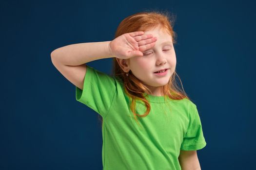 Funny photo of little redhead girl covering her eyes up from bright light or sun, blue background, green t-shirt