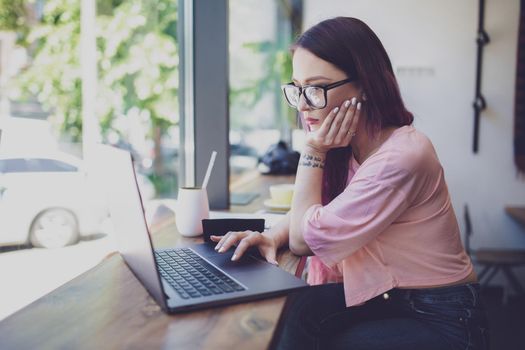 Side view of young businesswoman sitting at table in coffee shop. On table cup of coffee and laptop. In background white wall and window. Girl shopping online.