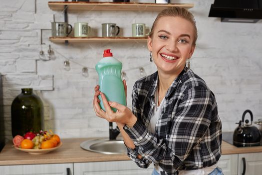 Young smiling housewife on kitchen. Housekeeper woman holding bottle with cleaner liquid. Bottle copy space